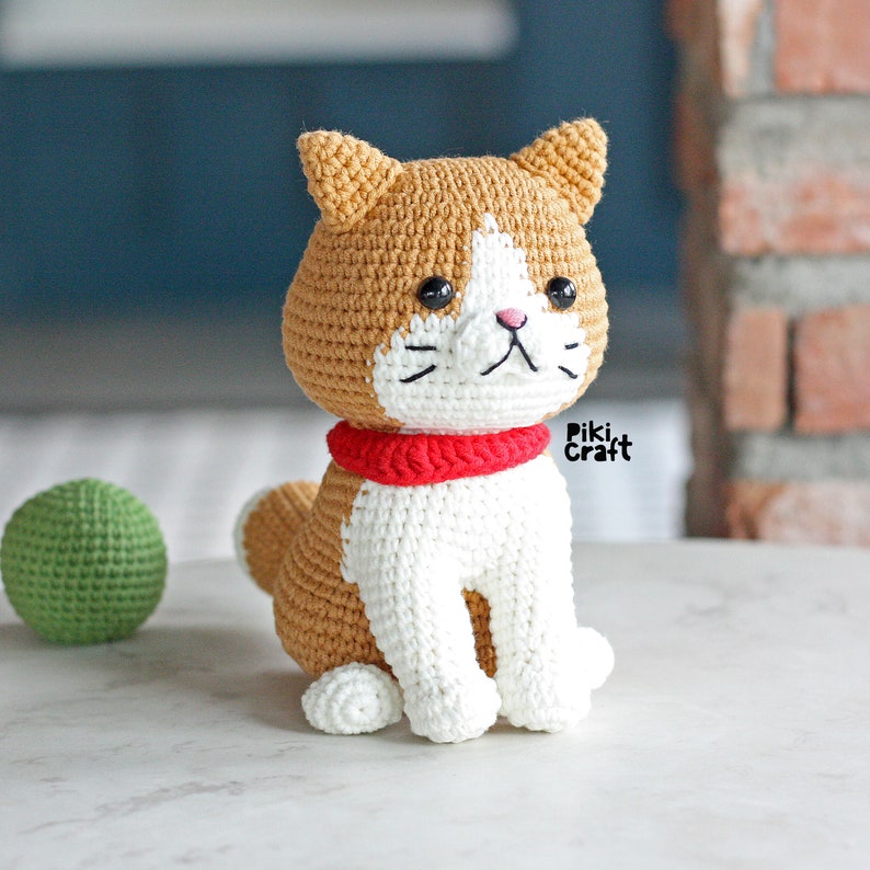 May include: A crocheted cat toy with brown and white fur, wearing a red scarf. The cat is sitting on a white surface.