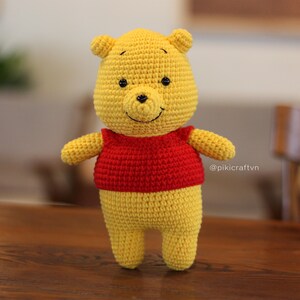 May include: A crocheted Winnie the Pooh plush toy wearing a red shirt. The toy is yellow with black eyes and a black nose. The toy is standing on a wooden surface.