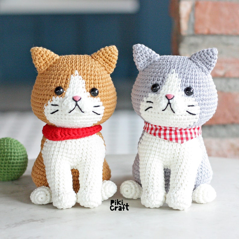 May include: Two crocheted cat toys, one orange and white with a red scarf, the other gray and white with a red and white checkered scarf.