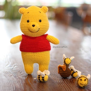 May include: A crocheted Winnie the Pooh plush toy wearing a red shirt stands on a wooden surface.  Three crocheted bee toys are also on the surface.  The bee toys are yellow and black with brown accents.  The image includes the text "@pikicraftvn".