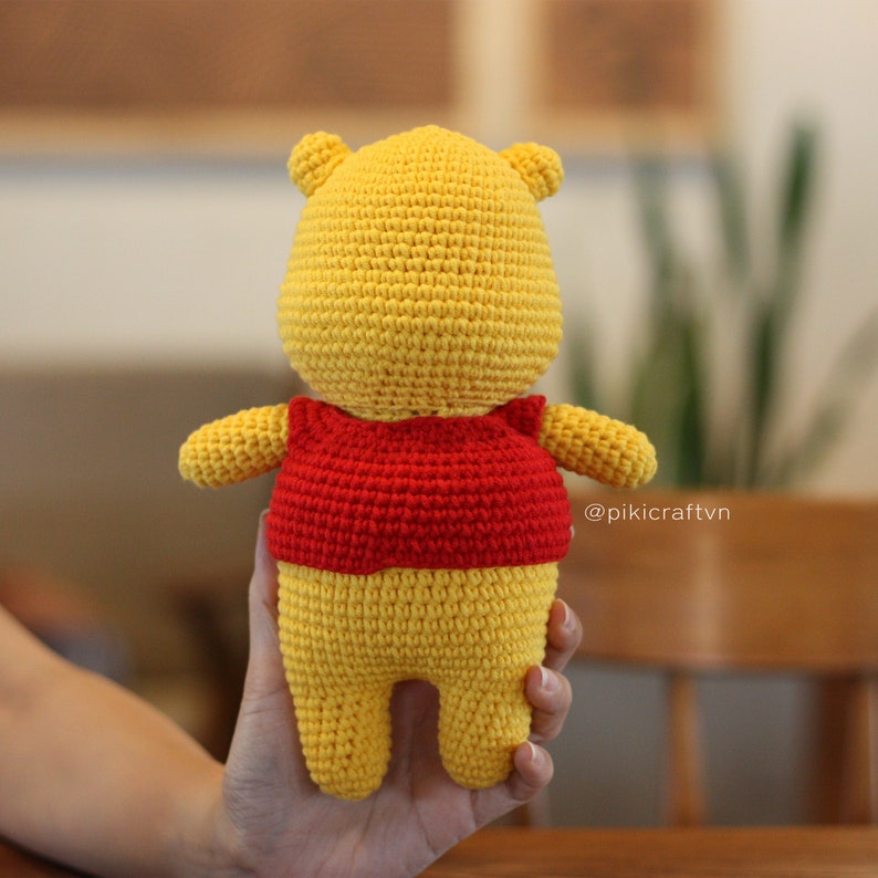 May include: A crocheted Winnie the Pooh plush toy wearing a red shirt. The toy is yellow and has a brown nose. The toy is being held by a hand.