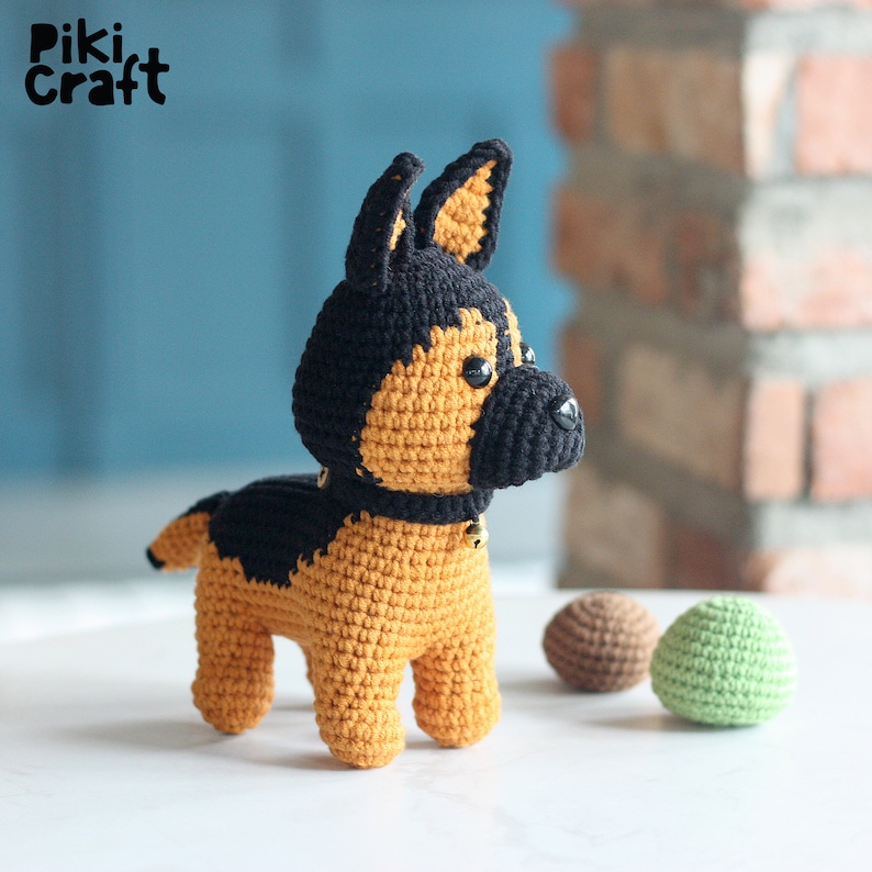 K&ouml;nnte beinhalten: Ein geh&auml;keltes Stofftier eines Deutschen Sch&auml;ferhundes mit schwarzem und braunem Fell. Der Hund tr&auml;gt ein schwarzes Halsband mit einer goldenen Glocke.