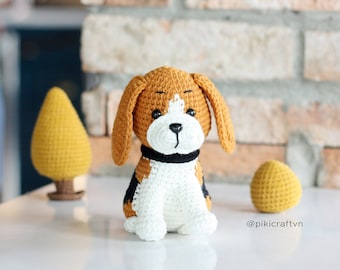 Puede incluir: Un juguete de peluche de perro beagle tejido a crochet con pelo marrón, blanco y negro. El perro está sentado con la cabeza ligeramente inclinada hacia un lado. El perro está sobre una superficie blanca con un pequeño árbol tejido a crochet y una pelota al lado.