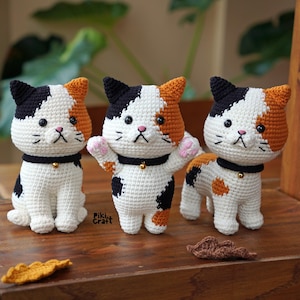 May include: Three handmade crochet cat figurines. Each cat has a white body with black and orange markings, black ears, and a black collar with a gold bell. The cats are displayed on a wooden surface.