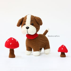 Puede incluir: Un perro de crochet marrón y blanco con una bufanda roja se encuentra de pie sobre una superficie blanca con dos setas de crochet rojas y blancas. El perro tiene una raya blanca en la cabeza y patas blancas. El texto '@pikicraftvn' está en el fondo.