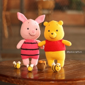May include: Two crocheted stuffed toys, a pink pig and a yellow bear, are sitting on a wooden table. The pig has black stripes and the bear has a red shirt. There are three small yellow crocheted bees and a small brown crocheted pot of honey on the table. The text '@pikicraftvn' is in the bottom right corner of the image.
