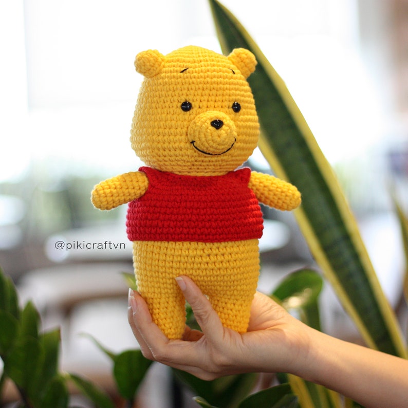 May include: A crocheted Winnie the Pooh plush toy wearing a red shirt. The toy is yellow and has a black nose and eyes. The toy is being held in a person's hand.