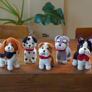 Op de afbeelding: Vijf gehaakte hondjes in verschillende rassen, waaronder een beagle, een dalmatiër, een bulldog, een schnauzer en een Franse bulldog. De knuffels staan allemaal op een houten oppervlak en hebben rood-witte halsbanden.