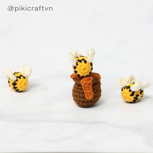 May include: Three crocheted bees, one on top of a brown crocheted honey pot, on a white surface. The bee on top of the pot has white wings. The text '@pikicraftvn' is in the upper left corner of the image.