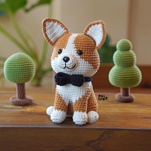 Może przedstawiać: Szydełkowa figurka psa corgi z brązowymi, białymi i czarnymi detalami. Pies ma czarną muszkę i siedzi wyprostowany. Po obu stronach psa znajdują się dwa małe zielone szydełkowe drzewka.