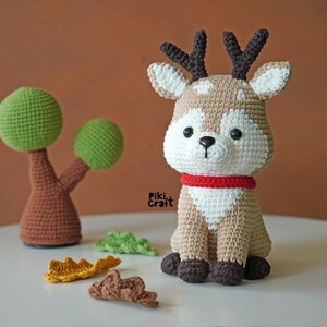May include: A crocheted reindeer toy with brown antlers, a white face, and a red collar. The reindeer is sitting on a white surface with brown and green crocheted leaves.