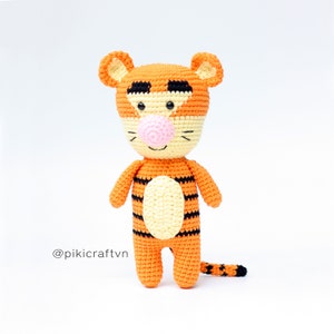 May include: A crocheted stuffed tiger toy with orange and black stripes, a yellow belly, and a pink nose. The toy has a friendly expression and is standing on a white background. The text '@pikicraftvn' is visible in the lower left corner of the image.