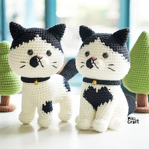 May include: Two crocheted black and white cat plushies with gold collars and bells. The cat on the left has a black spot on its leg. The cat on the right has a black heart on its chest. The cats are sitting on a white surface in front of two green crocheted trees.