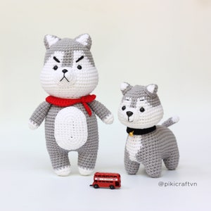 May include: Two crocheted husky dog plushies, one larger than the other. The larger husky is wearing a red scarf. The smaller husky is wearing a black collar with a gold bell. Both plushies have white accents on their faces and bodies. The plushies are standing on a white surface with a red toy bus in the foreground. The image includes the text "@pikicraftvn".