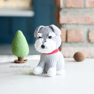 Könnte beinhalten: Ein grauer gehäkelter Zwergschnauzer mit weißen Akzenten und einem roten Halsband. Der Hund sitzt auf einer weißen Oberfläche.