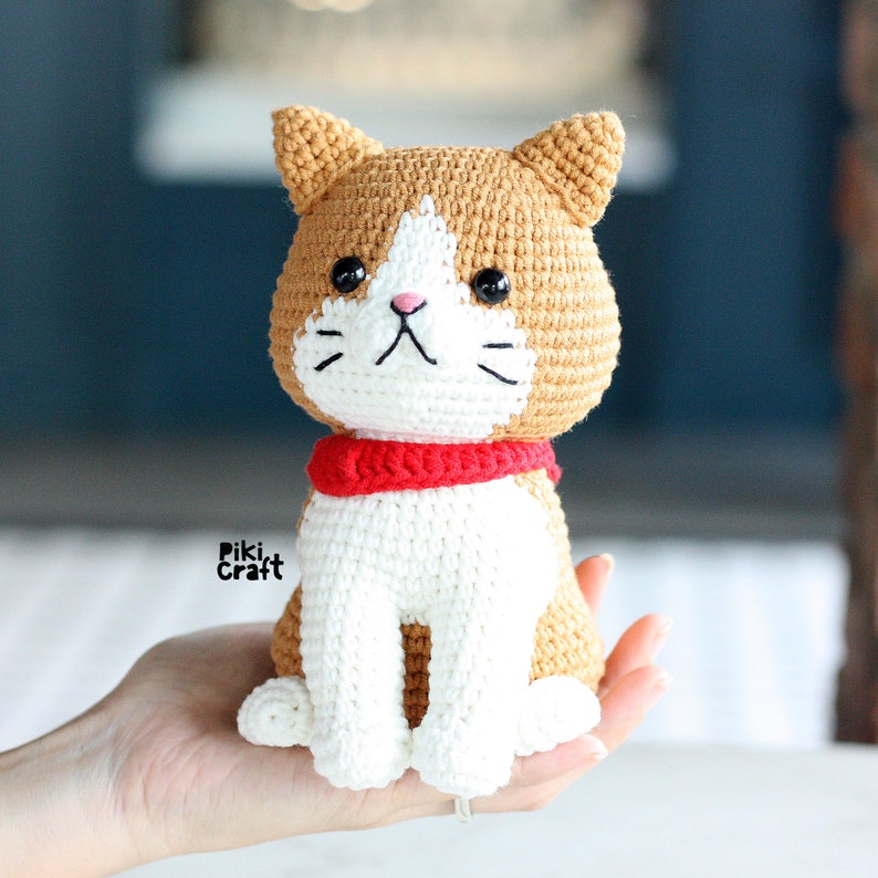 May include: A crocheted cat toy with white and brown fur, wearing a red scarf. The cat is sitting on a hand.