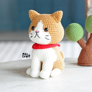 May include: A crocheted cat toy with white and brown fur, wearing a red scarf. The cat is sitting on a white surface.