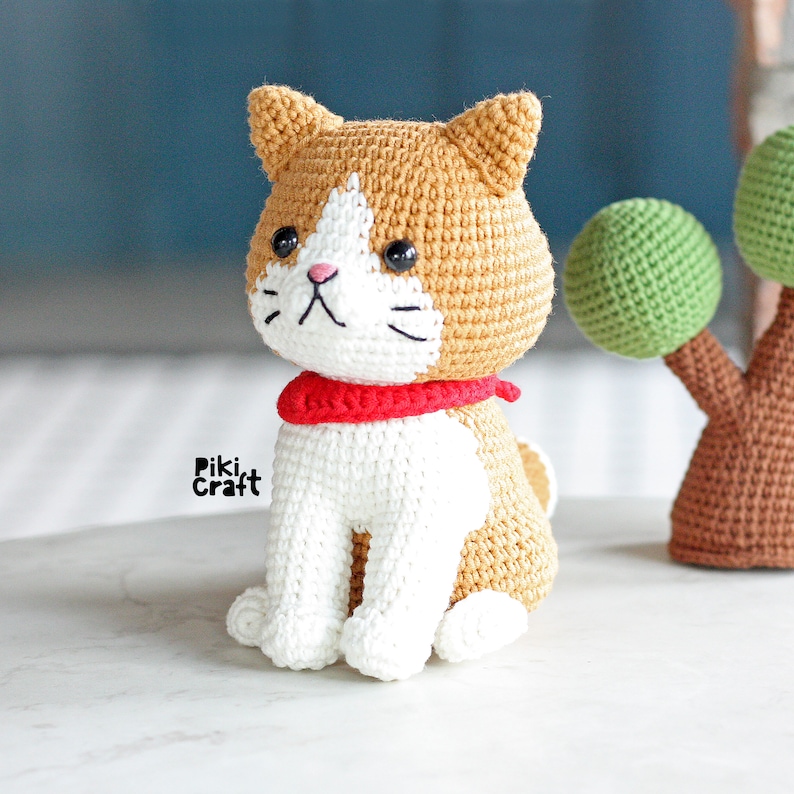 May include: A crocheted cat toy with white and brown fur, wearing a red scarf. The cat is sitting on a white surface.