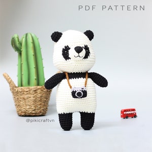 May include: A handmade crochet panda bear with black and white coloring, wearing a camera around its neck. The panda has black ears, arms, and legs. The text "PDF PATTERN" is at the top. A cactus and a red double-decker bus are in the background.