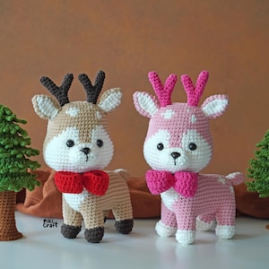 Peut inclure: Deux jouets en forme de rennes en crochet, l'un brun avec un nœud rouge et l'autre rose avec un nœud rose. Les jouets sont posés sur une surface brune avec des arbres verts derrière eux.