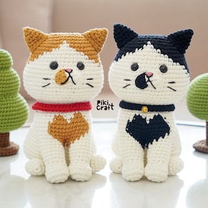 May include: Two crocheted cat plushies, one orange and white with a red collar, the other black and white with a black collar. Both are sitting on a white surface with green crocheted trees behind them.