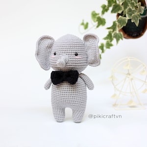 Puede incluir: Un elefante de ganchillo gris hecho a mano con una pajarita negra. El elefante tiene orejas grandes, una trompa y ojos negros pequeños. Las mejillas están ligeramente sonrojadas. El texto "@pikicraftvn" es visible en la parte inferior derecha.
