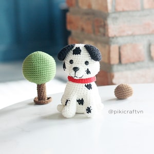Puede incluir: Un juguete de perro dálmata de crochet con manchas negras y un collar rojo. El perro está sentado sobre una superficie blanca con un árbol de crochet verde y una pelota marrón.