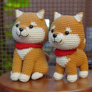 May include: Two crocheted Shiba Inu dog figurines. The dogs are tan, white, and brown with black eyes and noses. Each dog wears a red collar. One dog is sitting, and the other is standing. The figurines are on a wooden surface.
