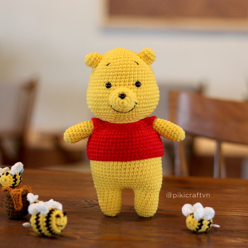 May include: A crocheted Winnie the Pooh plush toy wearing a red shirt, standing on a wooden table with three crocheted bees. The bees are yellow and black with white wings. The caption on the table reads "@pikicraftvn".