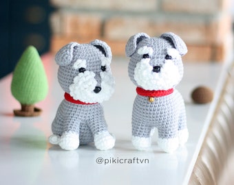 May include: Two gray crocheted miniature schnauzer dogs, one sitting and one standing, both wearing red collars with gold bells. The dogs are on a white surface with a brick wall in the background.