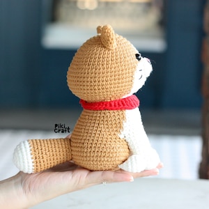 May include: A crocheted stuffed dog toy with a brown and white body and a red collar. The dog is sitting up with its head tilted up.