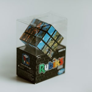 May include: A Rubik's Cube puzzle with a photo collage design is inside a clear plastic box. The box is sitting on a black box with the Rubik's Cube logo and the words "Check out the cool!" printed on it.