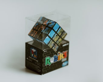 Puede incluir: Un rompecabezas Rubik's Cube con un diseño de collage de fotos está dentro de una caja de plástico transparente. La caja está sentada sobre una caja negra con el logotipo de Rubik's Cube y las palabras "Check out the cool!" impresas en ella.