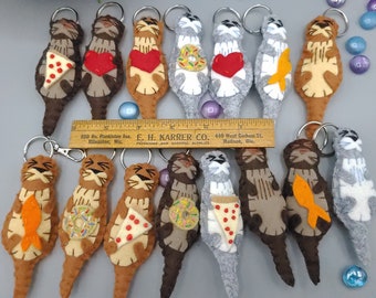 May include: A collection of 14 felt otter keychains. The otters are brown and gray and are holding various items, including a slice of pizza, a donut, a fish, and a heart. The keychains are attached to silver key rings.