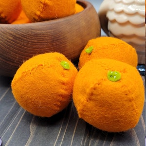 May include: A bowl of felt orange ornaments. The oranges are a bright orange color and have green felt stems. The bowl is made of wood and has a dark brown stain.