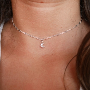 Peut inclure: Un collier en argent délicat avec un petit pendentif en forme de croissant de lune. La chaîne est une fine conception à maillons, et le collier est porté autour du cou d'une personne. Le bijou est simple et élégant.