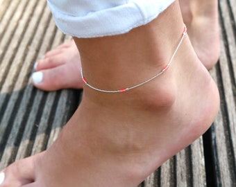 May include: A silver anklet with small red beads. The anklet is on a person's ankle.