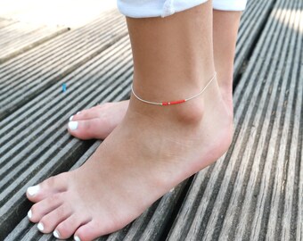 May include: A silver anklet with a small red accent on a wooden surface.