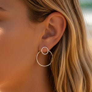 以下が含まれることがあります： 小さな円が大きなフープに付いたシルバーのイヤリングのクローズアップ。イヤリングは人の耳に着用され、金髪が見えます。背景は、遠くに海が見えるビーチのシーンです。