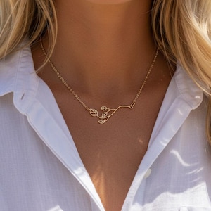 May include: A gold-coloured necklace with a branch and leaf design. The necklace is worn against a white collared shirt, with a beach scene in the background. The necklace is a simple, elegant piece of jewellery, suitable for everyday wear.