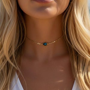 May include: A fine gold choker necklace with a teardrop-shaped, dark teal gemstone pendant. The necklace is worn around the neck, with the pendant at the base of the throat. The background is blurred, suggesting an outdoor setting.