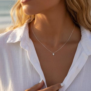 Peut inclure: Un délicat collier en argent avec un petit pendentif en forme de cœur. Le collier est porté sur une chemise blanche. L'image est un gros plan, mettant l'accent sur le collier et le décolleté.