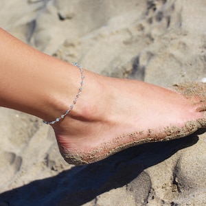 Peut inclure: Un bracelet de cheville argenté avec de petites perles transparentes sur la cheville d'une femme. La cheville repose sur du sable.