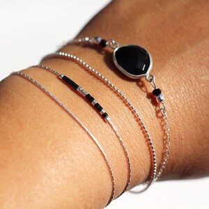 Pode incluir: Um conjunto de quatro pulseiras de prata, uma com contas pretas, outra com uma pedra preciosa preta e duas correntes finas de prata.