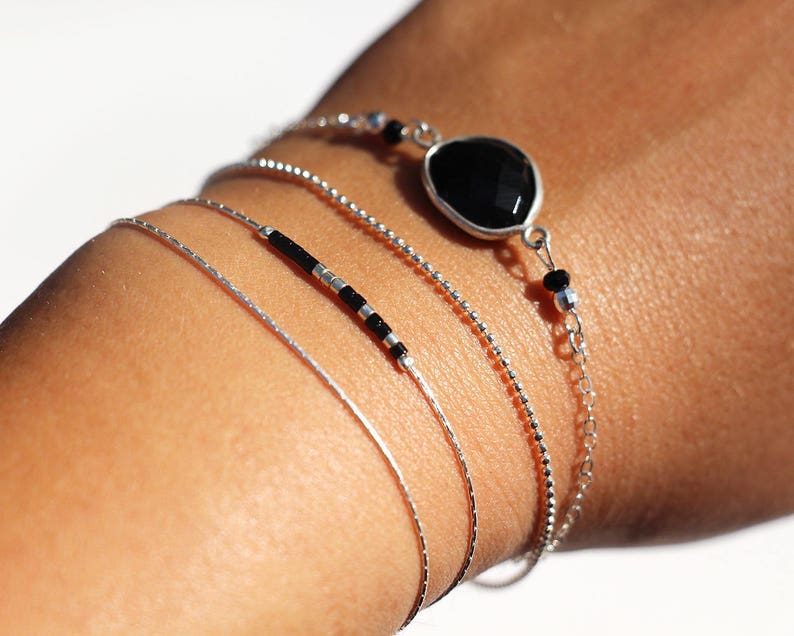 Pode incluir: Um conjunto de quatro pulseiras de prata, uma com contas pretas, outra com uma pedra preciosa preta e duas correntes finas de prata.