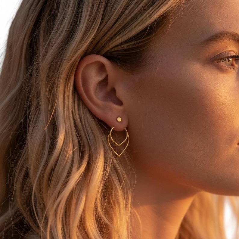 May include: A close-up of a person's ear adorned with two gold earrings. The upper earring is a small, round stud, while the lower earring features a double teardrop design. The earrings are set against a backdrop of blonde hair and warm sunlight.