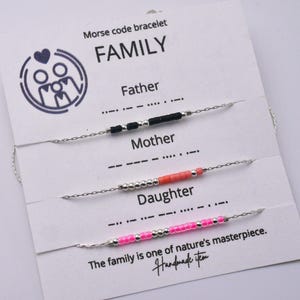 Puede incluir: Una tarjeta blanca muestra tres pulseras con código Morse para "Padre", "Madre" e "Hija". Las pulseras tienen cadenas plateadas y cuentas negras, coral y rosas. La tarjeta dice "FAMILIA" y "La familia es una obra maestra de la naturaleza."