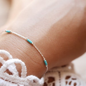 Puede incluir: Un delicado brazalete de cadena de plata con pequeñas cuentas turquesas. El brazalete está en la muñeca de una persona.