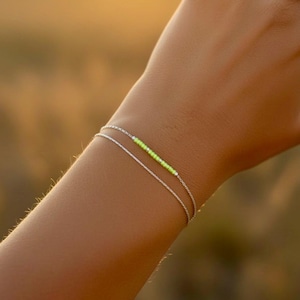 May include: A delicate silver bracelet with two fine chains. The top chain features a row of small, bright neon green beads. The bracelet is shown on a person's arm, with a blurred background.