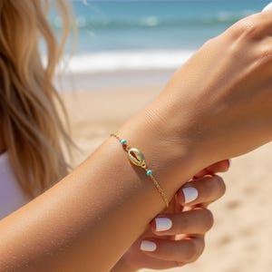Op de afbeelding: Een goudkleurige armband met een schelpenbedel en kleine turquoise kralen. De fijne kettingarmband wordt om een pols gedragen, met een strand en zee op de achtergrond. De armband is een sieraad.